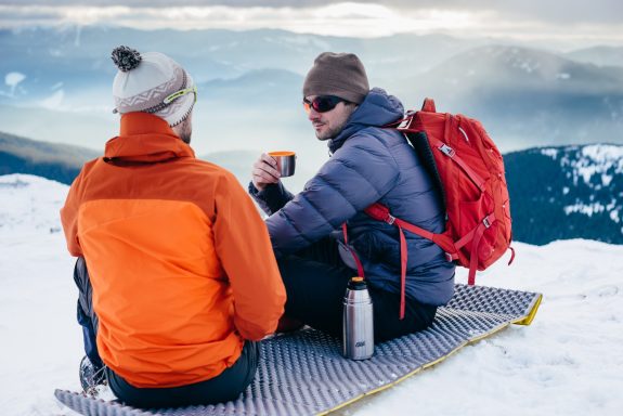 Zwei Männer in bergiger Schneelandschaft genießen ein heißes Getränk aus einer Esbit Thermoskanne.