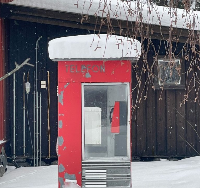 Rote Telefonzelle mit Schnee bedeckt, vor einem rustikalen Holzgebäude.