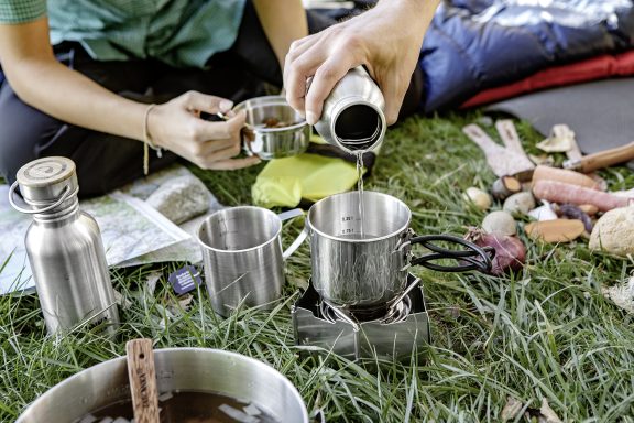 Zwei Leute bereiten ihre  Campingmalzeit mit Tatonka Edelstahl Trinkflaschen, Edelstahl Tassen und Tatonka Kocher mit Windschutz zu.